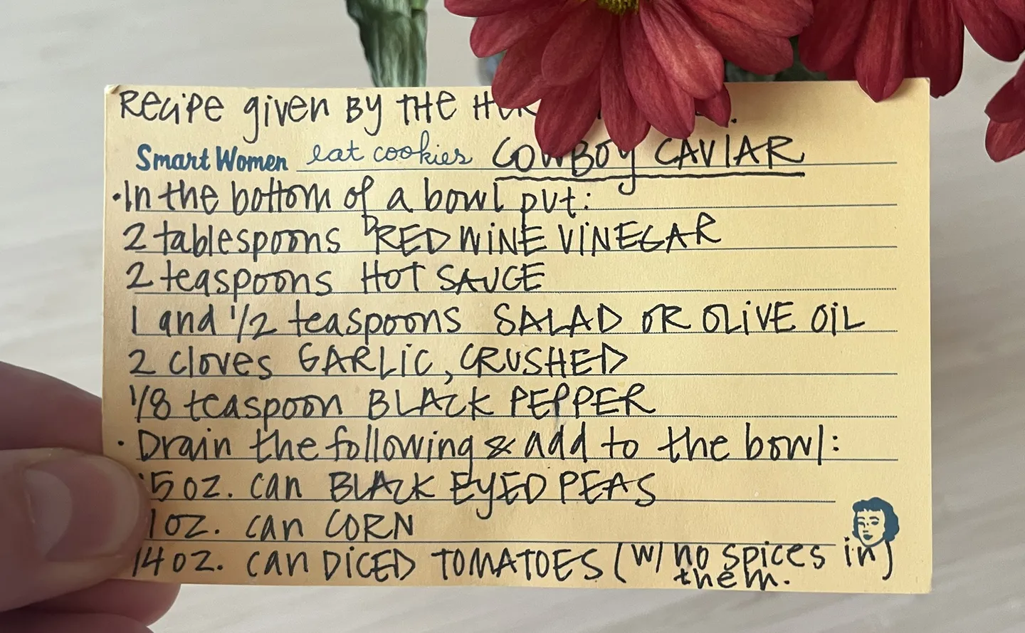 A handwritten Cowboy Caviar recipe card held up beside red flowers, listing red wine vinegar, hot sauce, oil, garlic, pepper, black-eyed peas, corn and diced tomatoes